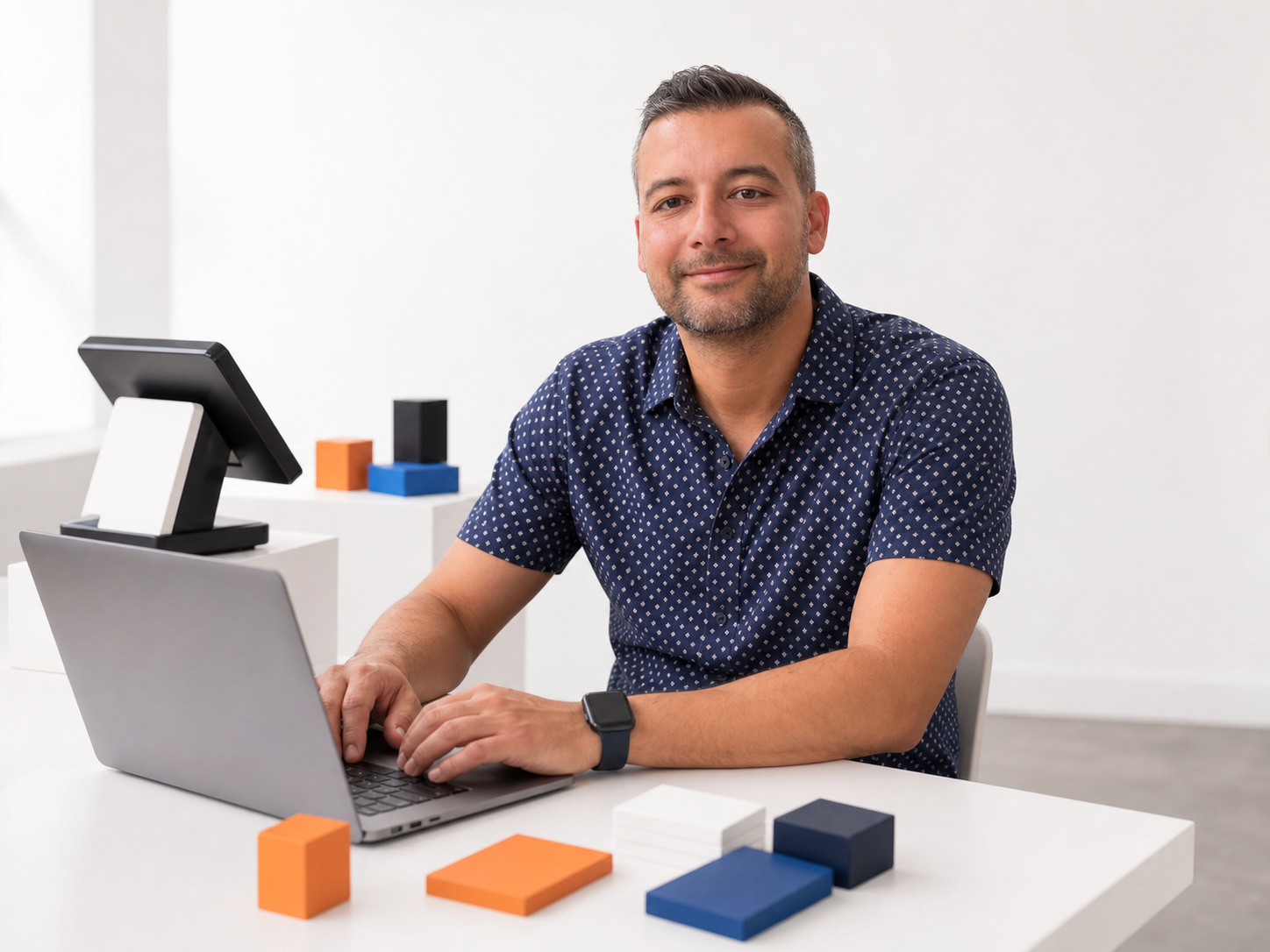 Jalil smiling at a white desk with a MacBook, checkout register, and abstract strategy blocks
